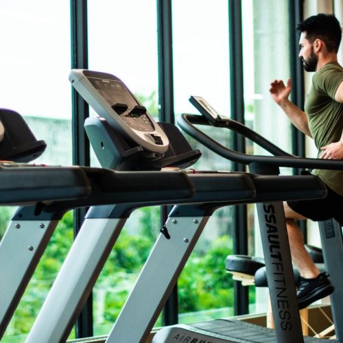 Young man workouts on treadmill in modern gym with large windows and natural light.